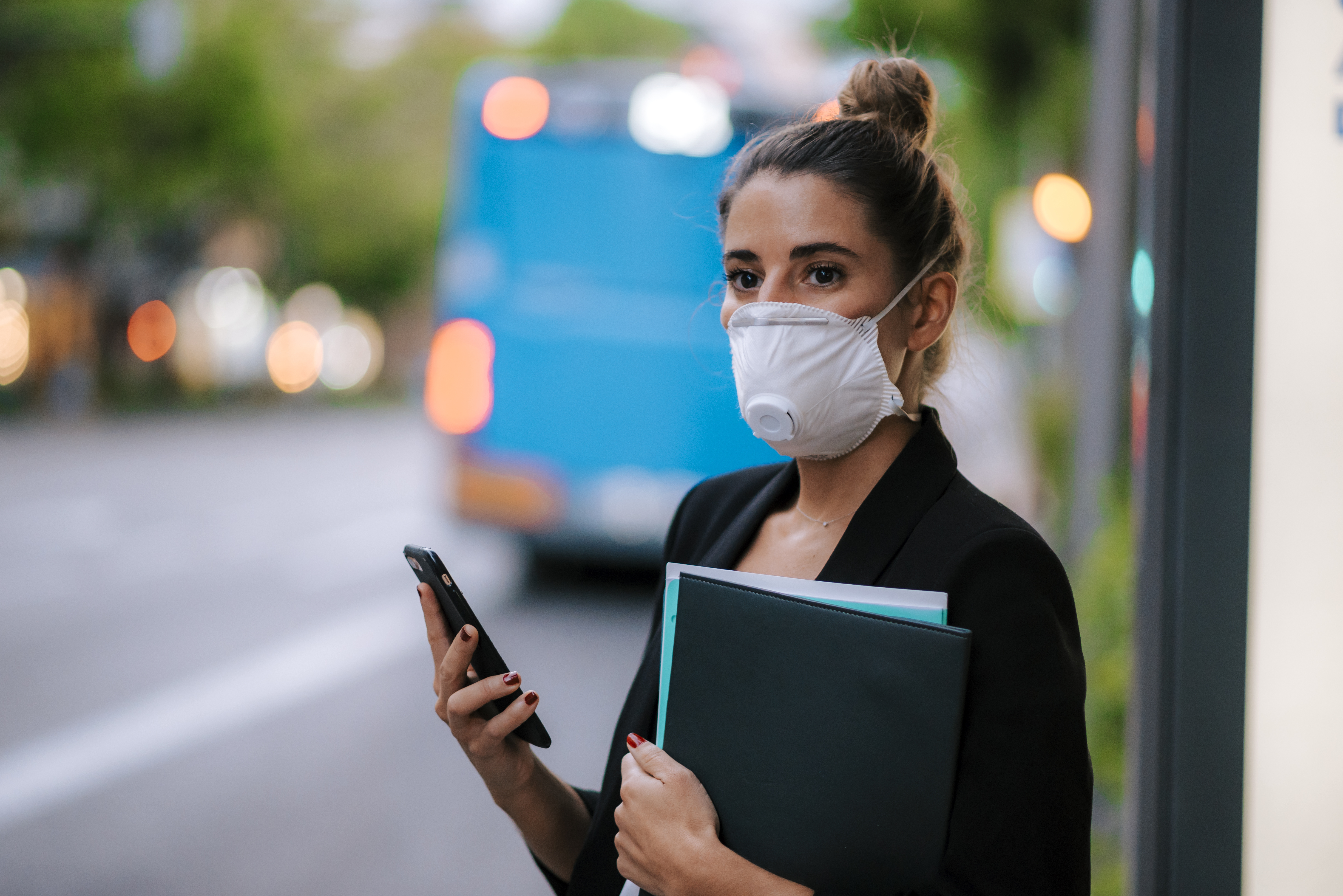 woman with mask holdingp hone and waiting for bus